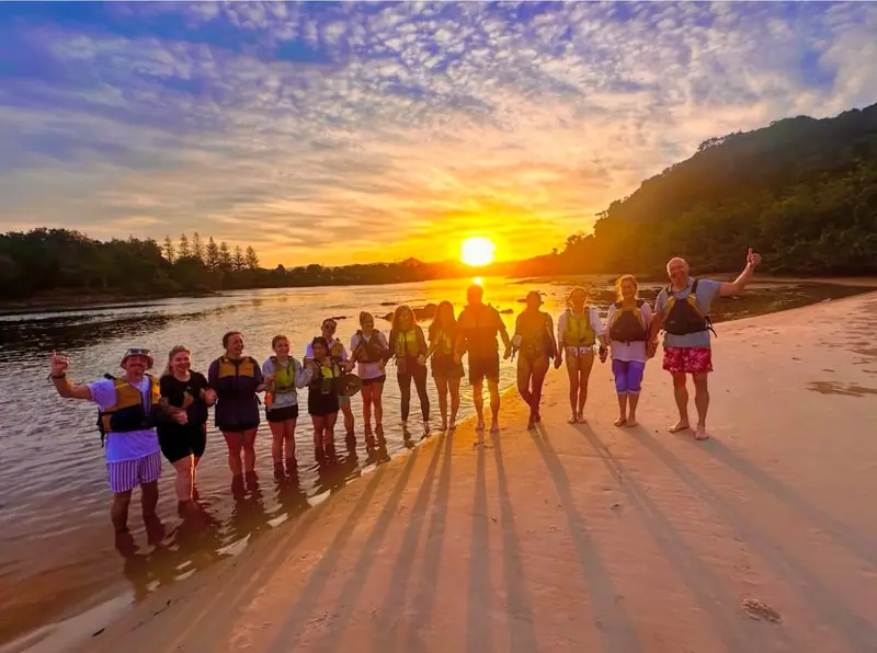 A happy tour group in life jackets holding hands on the riverbank at sunset
